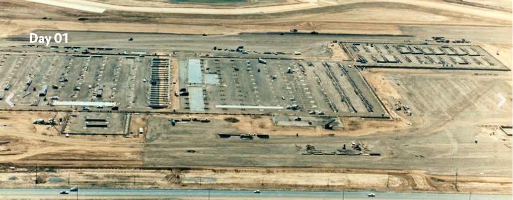 Aerial view of a large construction site labeled “Day 01,” showing cleared land, building foundations, structural framing, and construction equipment spread across a desert landscape.